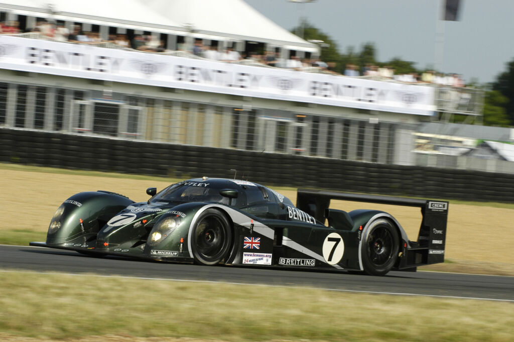 A 2003 Bentley Speed 8 being driven at speed past a Bentley liveried spectator stand