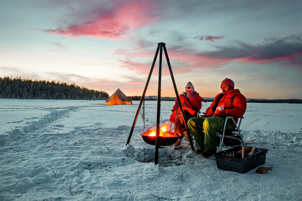 A couple cooking a meal sat in chairs on top of the snow