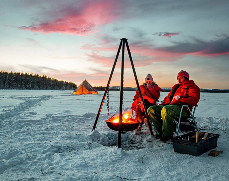 A couple cooking a meal sat in chairs on top of the snow