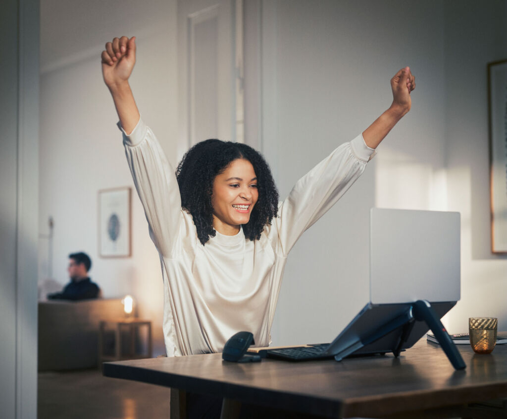 A woman in front of her computer, celebrating with her hands in the air