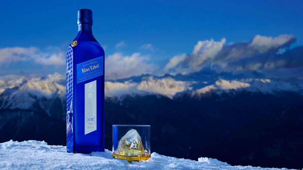 A bottle of Johnnie Walker Blue Label Ice Chalet and a full glass on top of snow atop a mountain